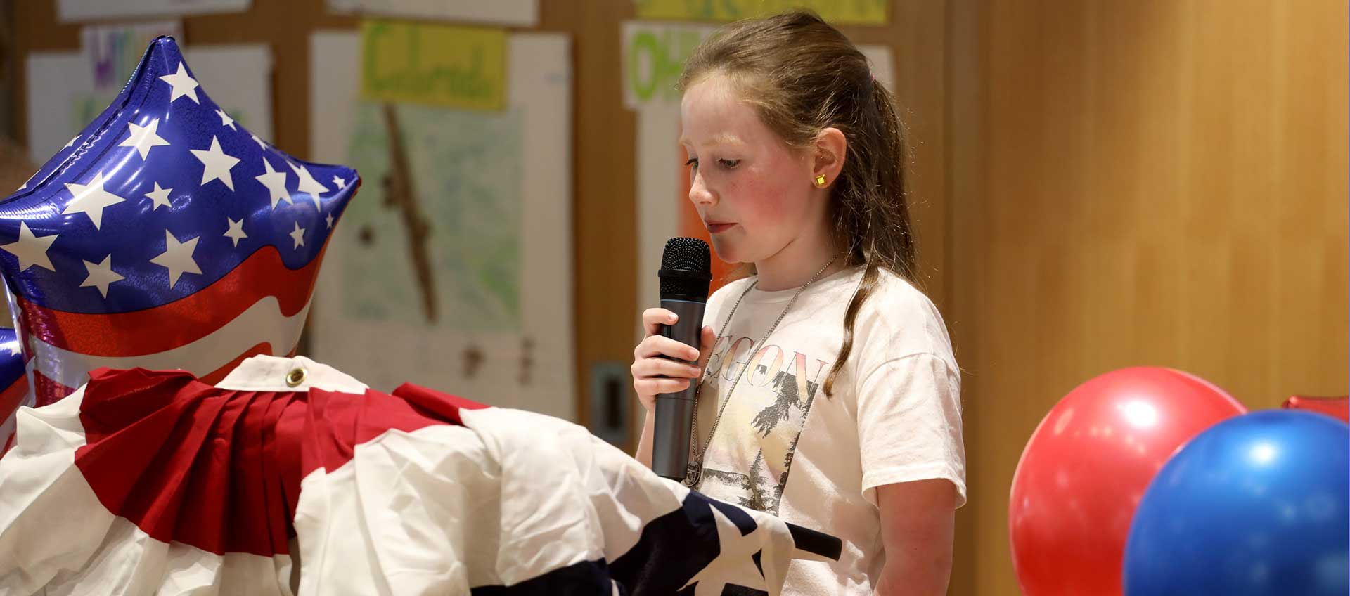 A student stands holding a microphone surrounded by red, white, and blue balloons