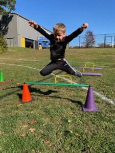 A pre-k student joyfully jumps over an obstacle in PE class