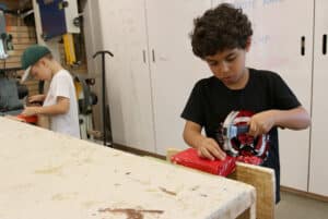 A first grader hammers their happy block in the woodworking studio