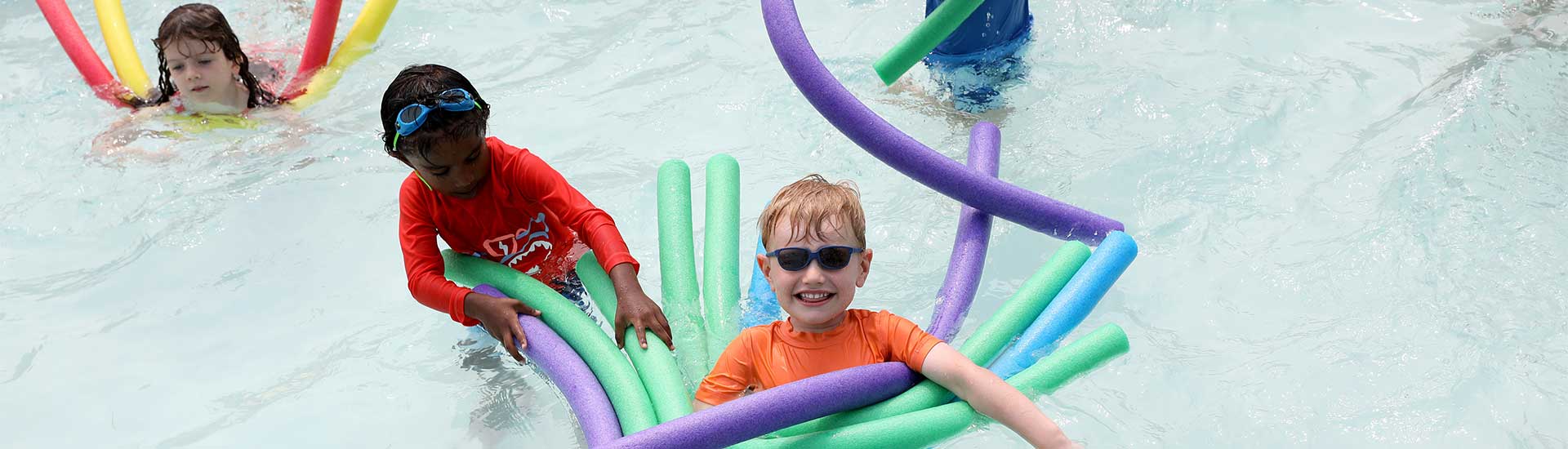 Three campers in the pool floating with colorful noodles