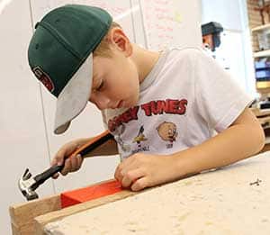 A student wearing cap works on hammering an orange piece of wood